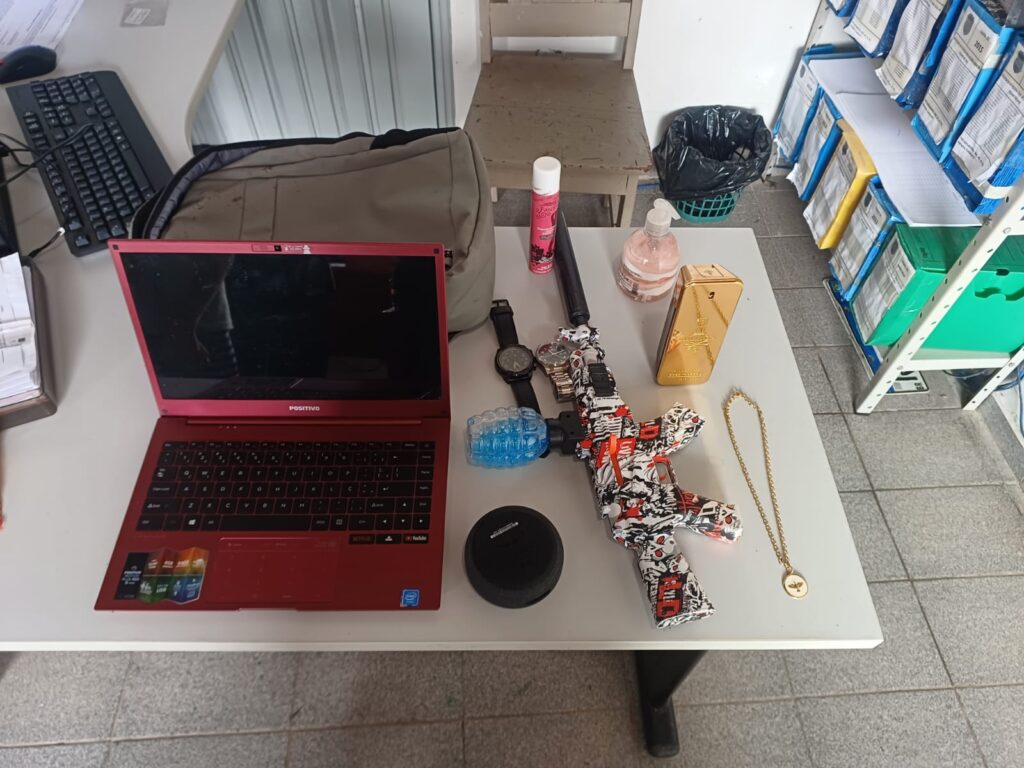 Objetos apreendidos: laptop vermelho, réplica de fuzil, relógios, perfume e joia sobre mesa branca.