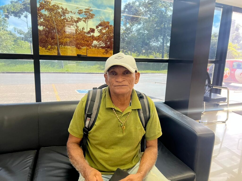 Homem de meia-idade sentado em um sofá preto, usando boné, camisa polo verde e mochila.
