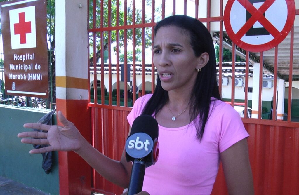 Mulher com camiseta rosa falando em microfone da SBT, expressão séria.