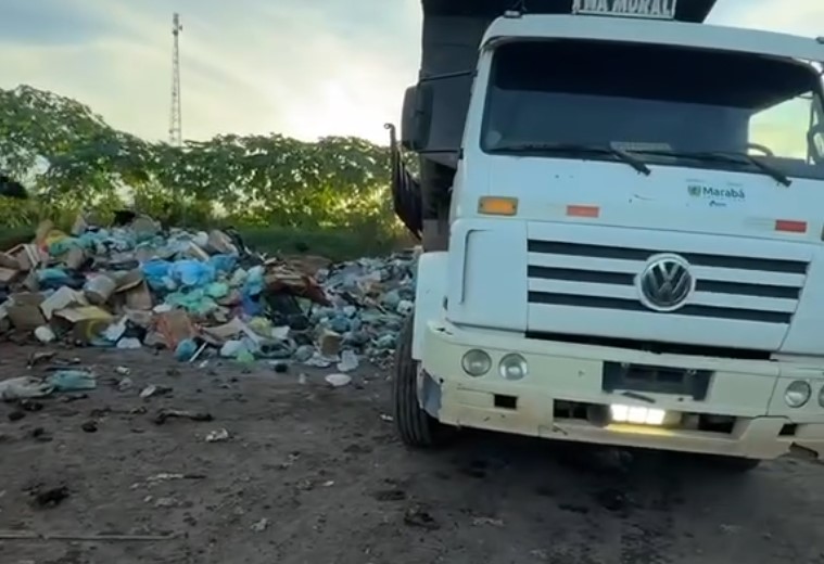 Caminhão de lixo branco da Volkswagen ao lado de uma grande pilha de resíduos em um terreno de terra.