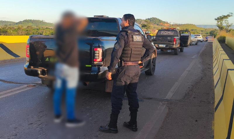Oficial da ROMU ao lado de veículo e pessoa borrada em operação policial.