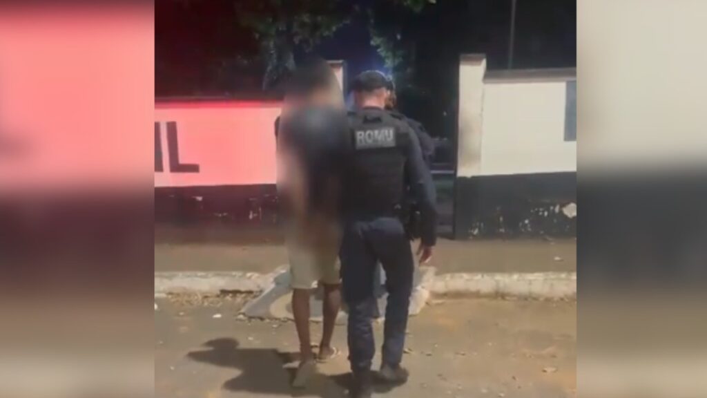 Policial da ROMU escoltando um indivíduo com o rosto desfocado em ambiente noturno.