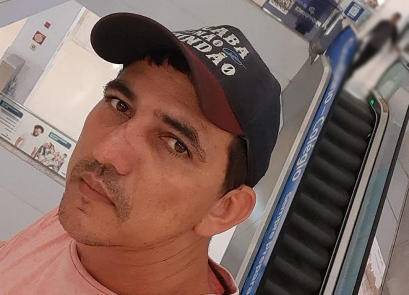 Homem com boné escuro e bigode olhando para a câmera, com fundo de escada rolante.
