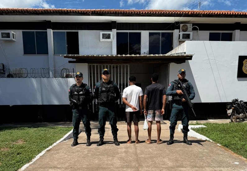 Policiais militares e dois indivíduos detidos em frente a uma delegacia.