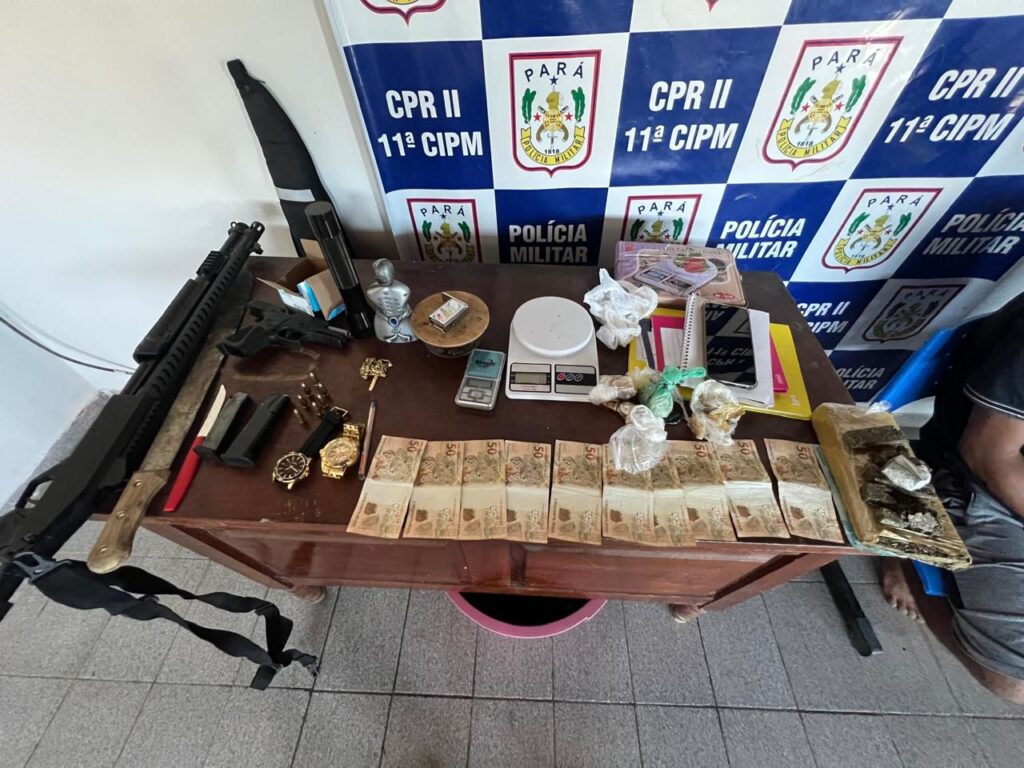 Visão aérea de uma mesa cheia de itens apreendidos pela polícia, incluindo armas de fogo, pilhas de dinheiro, drogas e balanças.