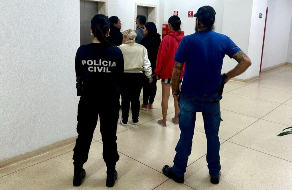 Policial civil e grupo de pessoas, algumas algemadas, em corredor com elevadores.