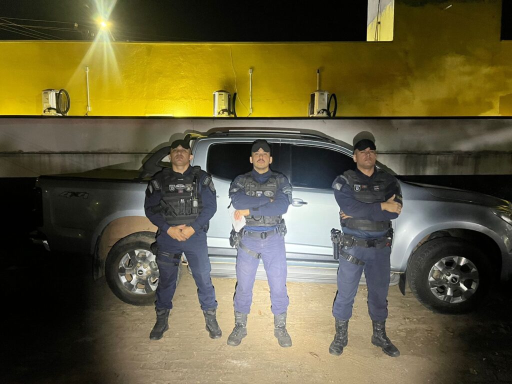 Três policiais ou seguranças uniformizados com coletes táticos em frente a uma picape prateada à noite.