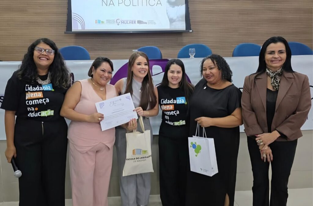 Grupo de mulheres sorridentes em palco durante evento, uma delas segurando um certificado.