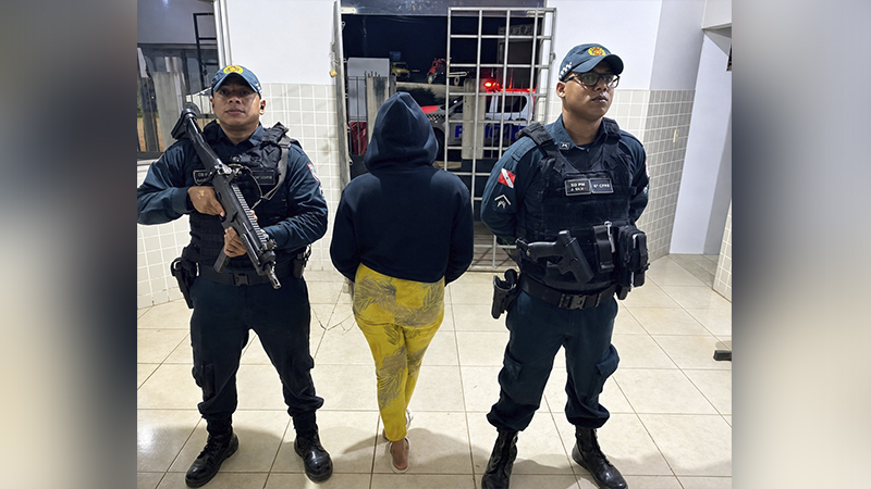 Dois policiais fardados com armas flanqueando uma pessoa de costas, em ambiente interno com grades e luzes de viatura ao fundo.