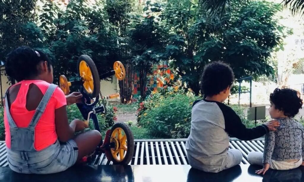 Crianças brincando com bicicleta azul e amarela em jardim verde.