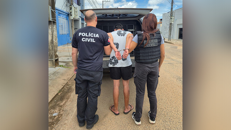 Homem algemado sendo escoltado por dois policiais civis em via pública.