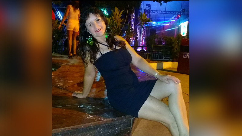 Mulher sorrindo em vestido preto sentada em ambiente noturno com luzes coloridas.