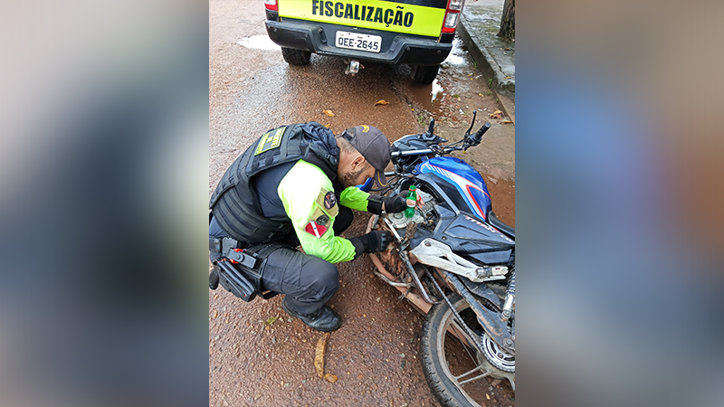 Agente de fiscalização ajoelhado inspecionando uma motocicleta azul e branca.