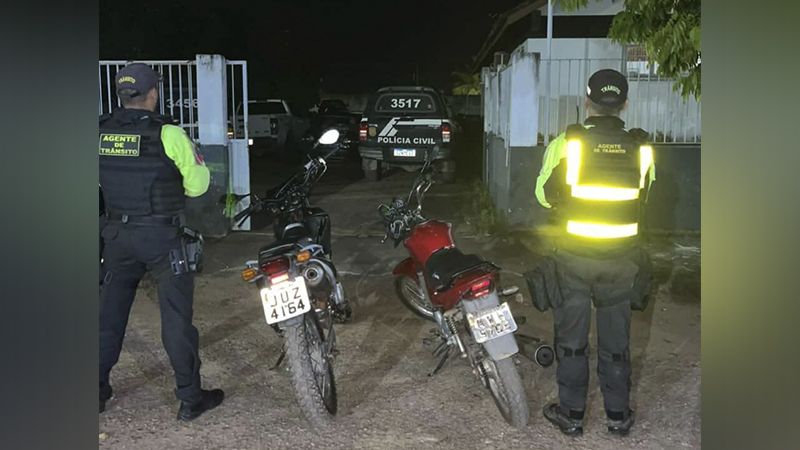 Dois agentes de trânsito em uniforme refletivo inspecionando duas motocicletas em uma operação noturna, com um veículo da Polícia Civil ao fundo.