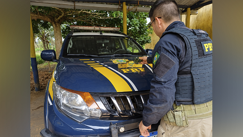 Policial da PRF ao lado de viatura azul com emblema no capô.