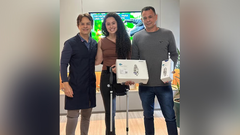 Três pessoas sorrindo: uma mulher com muletas e uma caixa Ottobock, um profissional de saúde e outro homem.