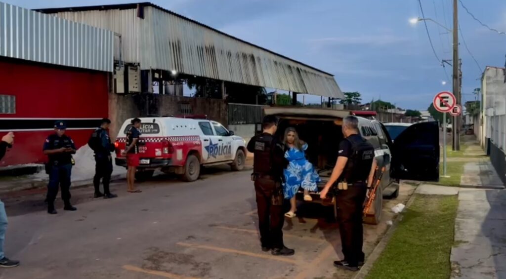 Mulher em vestido azul sendo conduzida por policiais em uma viatura.