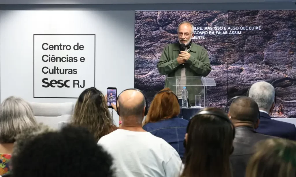Homem de barba palestrando no palco do Centro de Ciências e Culturas Sesc RJ para uma plateia atenta.