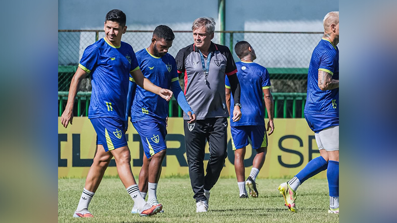 Treinador e jogadores de futebol em uniforme azul e amarelo durante um treino no gramado.