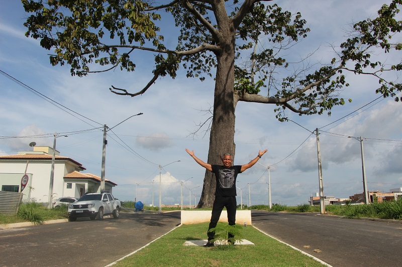 Homem sorrindo com braços abertos sob uma grande árvore em uma rua residencial pavimentada.