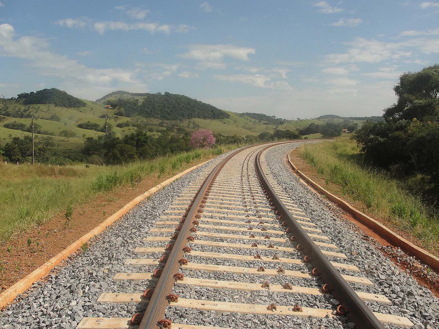Trilhos de trem em curva atravessando uma paisagem rural montanhosa em dia ensolarado