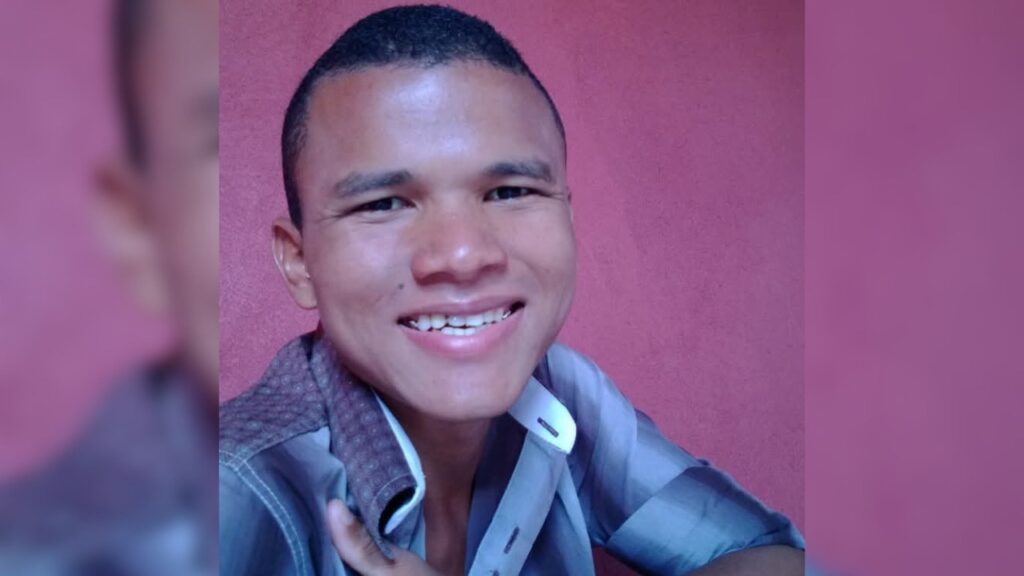 Jovem sorridente com camisa listrada em frente a parede rosa.
