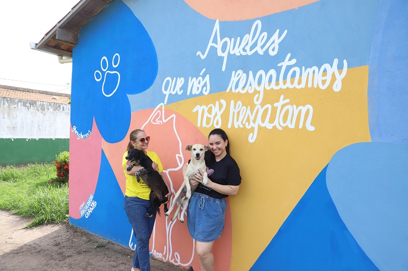 Duas mulheres sorridentes segurando cachorros em frente a um mural colorido com a frase 'Aqueles que nós resgatamos nos resgatam'.