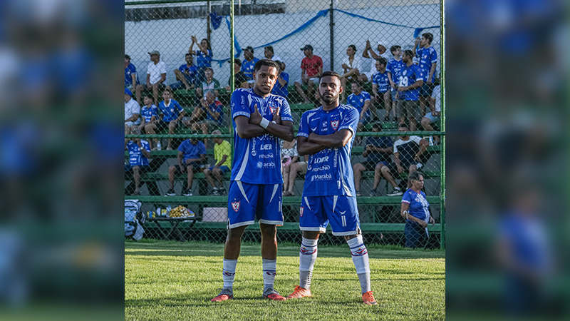 Dois jogadores de futebol em uniformes azuis posando em campo com torcida ao fundo.
