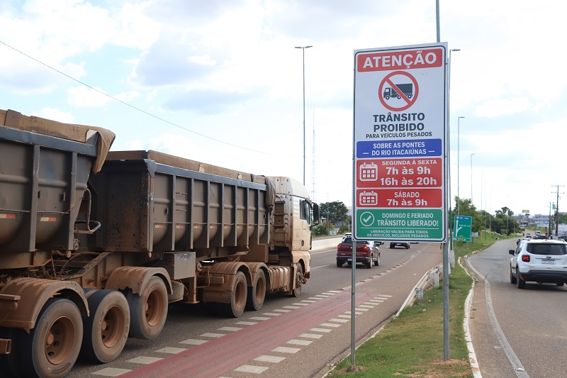 Um caminhão caçamba sujo passando por uma placa que proíbe o trânsito de veículos pesados em horários específicos.