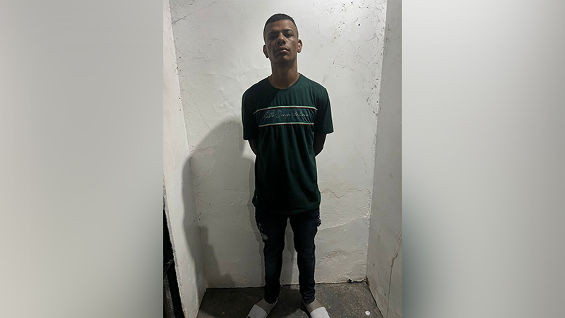 Homem jovem com camiseta verde e calça escura, parado em ambiente simples, olhando para frente.