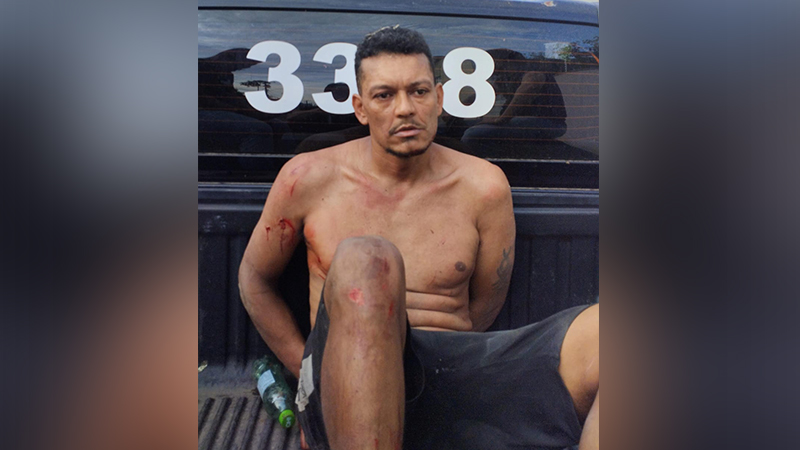 Homem sem camisa com ferimentos e algemado em parte traseira de veículo policial.