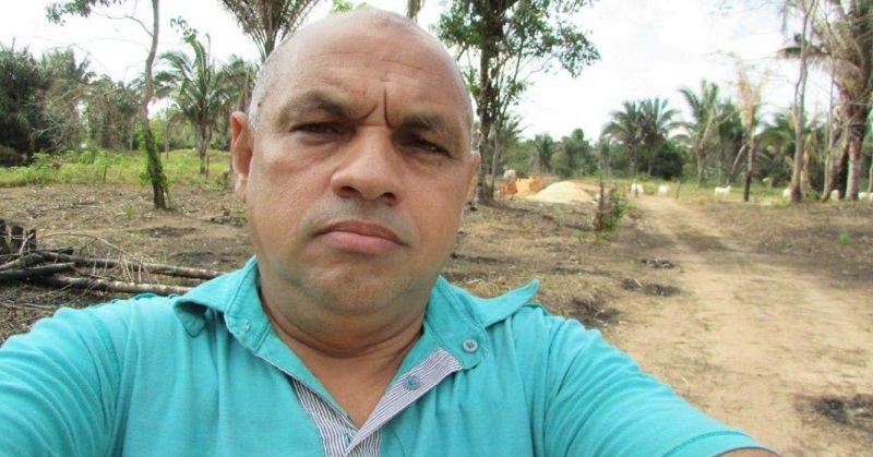 Homem careca de camisa azul-turquesa fazendo selfie em área rural.