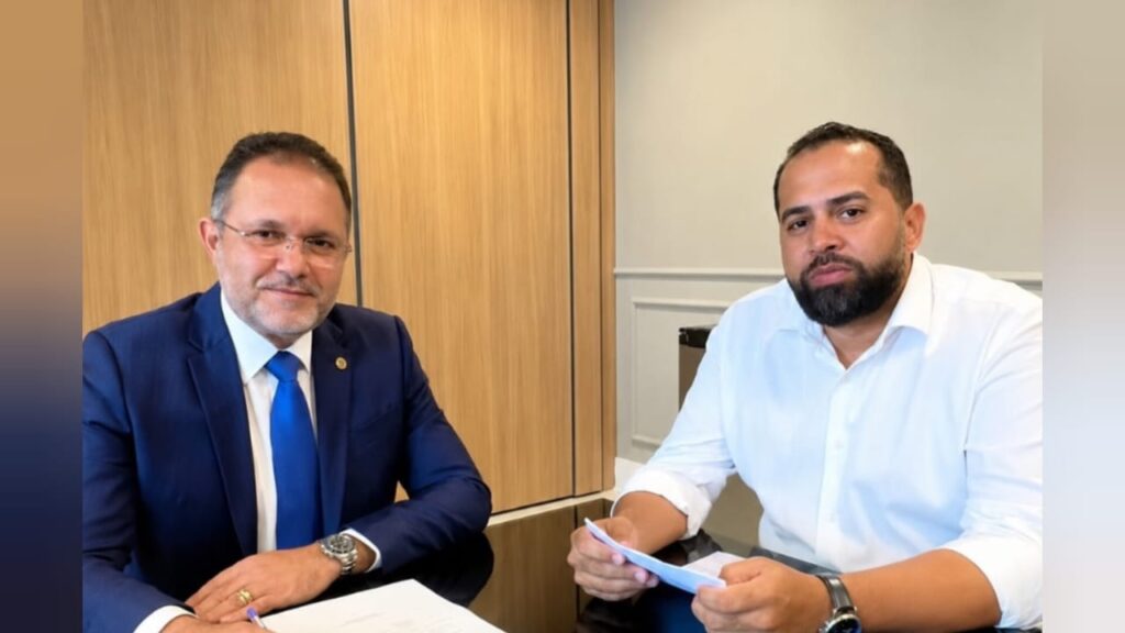 Dois homens sentados em mesa de escritório, um de terno azul e outro de camisa branca, com papéis em mãos.
