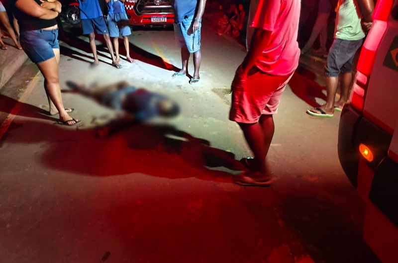 Corpo desfocado de uma pessoa caída na rua, cercado por curiosos e luzes vermelhas de emergência.