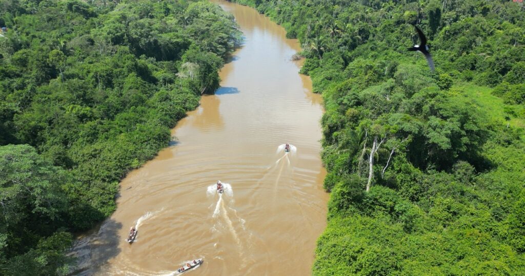 Vista aérea de um rio marrom serpenteando por uma floresta amazônica densa, com vários barcos motorizados navegando.