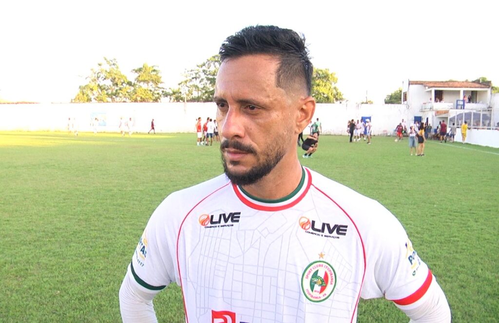 Homem com barba e cabelo cortado vestindo camisa de futebol branca em campo.