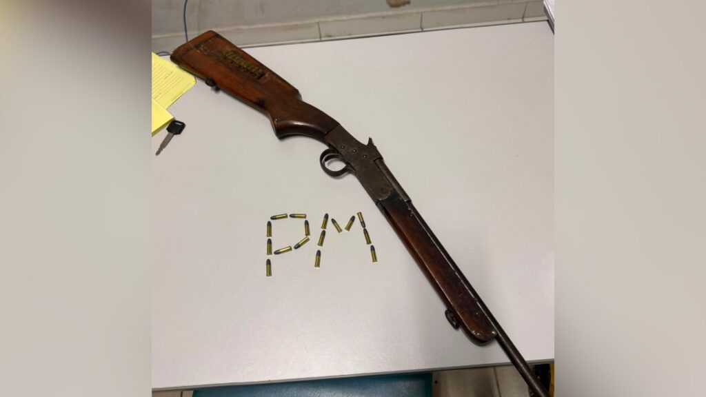 Espingarda de madeira e metal com munições formando 'PM' sobre uma mesa branca.