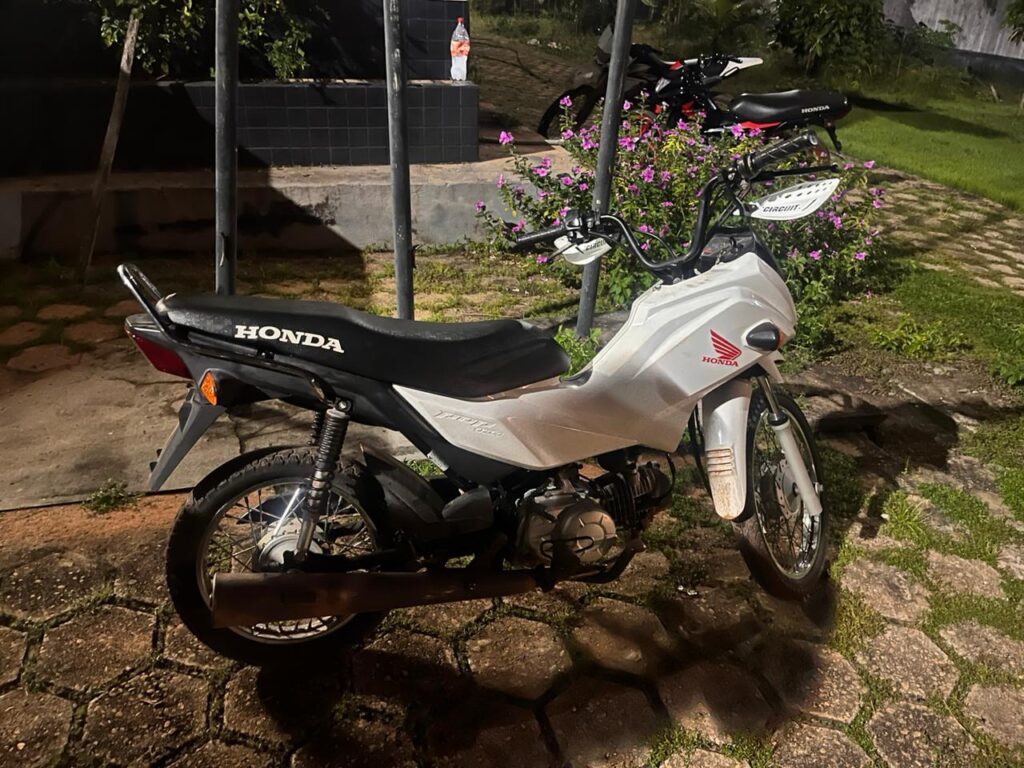 Moto Honda Pop branca estacionada em calçada de paralelepípedos perto de arbustos com flores.