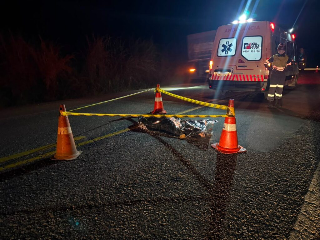 Corpo coberto no asfalto com fita de isolamento, cones, ambulância e equipe de emergência à noite.