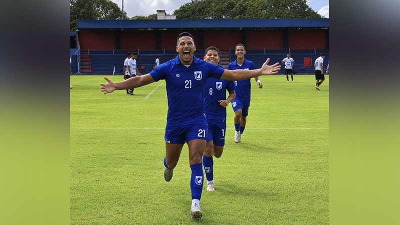 Jogador de futebol em uniforme azul vibrando com os braços abertos em um campo de grama, com colegas atrás.