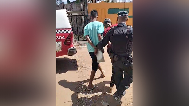 Homem algemado sendo escoltado por policial militar perto de viatura.