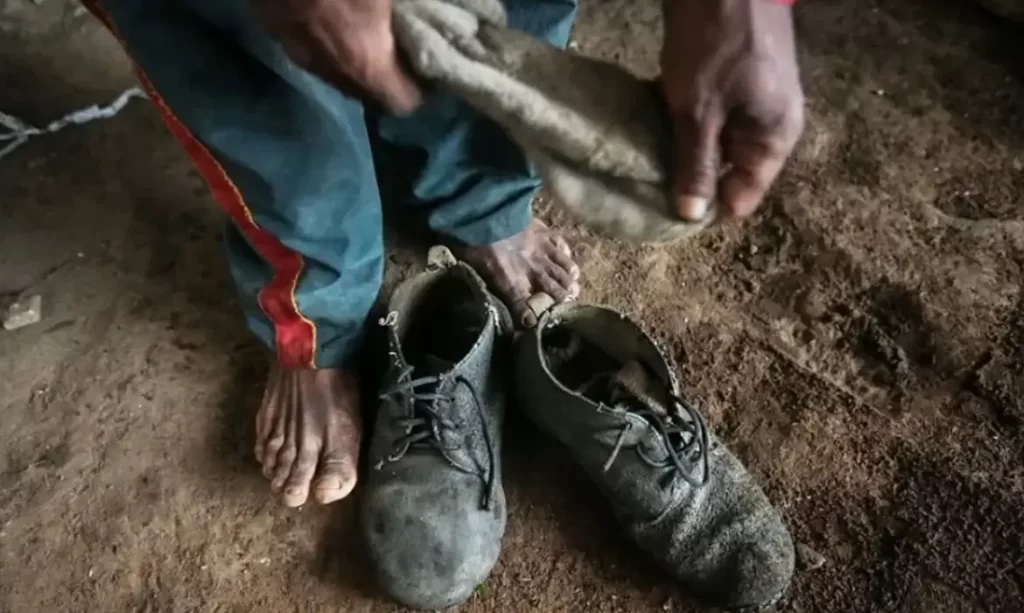 Pessoa descalça se preparando para calçar sapatos velhos e empoeirados sobre chão de terra.