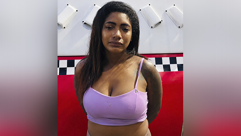 Mulher com cabelos escuros e top lilás em frente a um fundo vermelho e quadriculado.