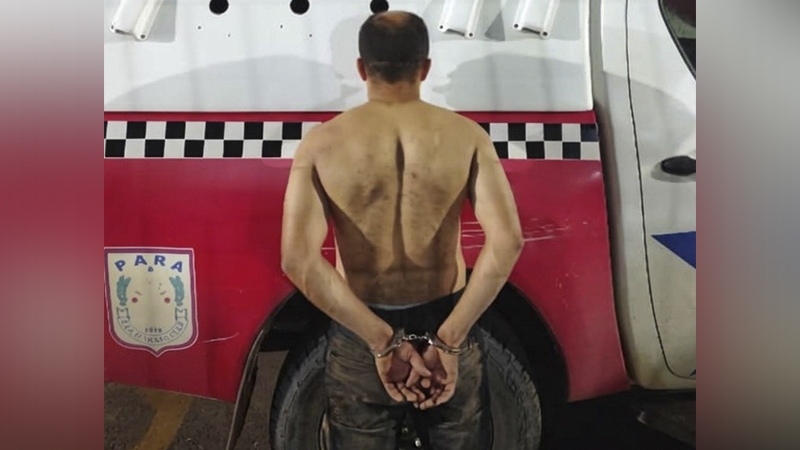 Homem algemado de costas, sem camisa, em frente a veículo policial.