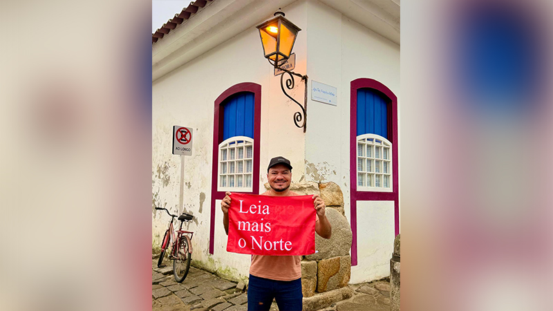 Homem sorridente segurando uma bandeira vermelha com a frase Leia mais o Norte em uma rua histórica.