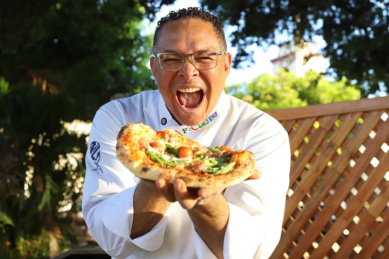 Chef de cozinha sorrindo entusiasticamente segurando uma pizza artesanal em um ambiente externo.