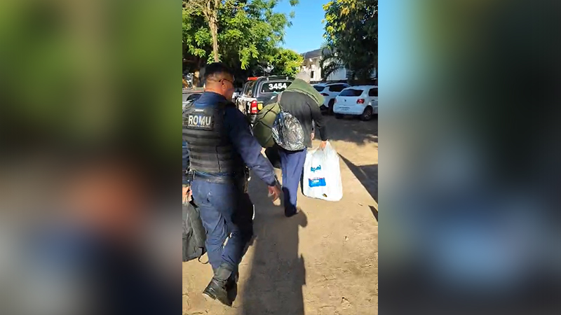 Agente da ROMU acompanhando um indivíduo que carrega bolsas em área externa durante o dia.