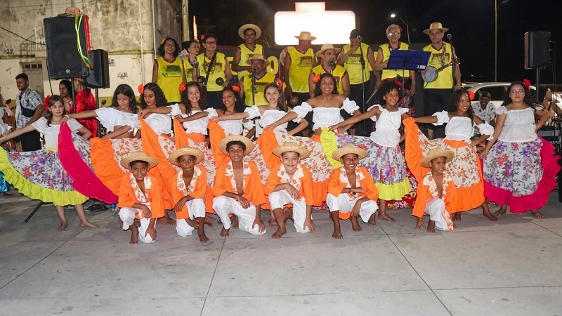 Grupo de crianças vestidas com trajes típicos de festa junina posando para foto em um palco ao ar livre.