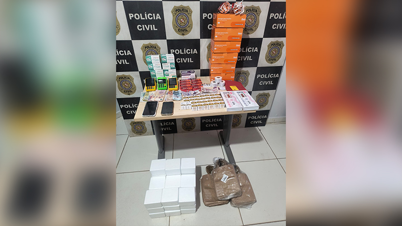 Mesa com itens apreendidos pela Polícia Civil incluindo caixas de medicamentos, máquinas de cartão e dinheiro.