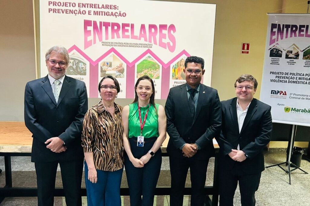 Grupo de cinco profissionais posando em frente ao painel do Projeto Entrelares de prevenção à violência doméstica.